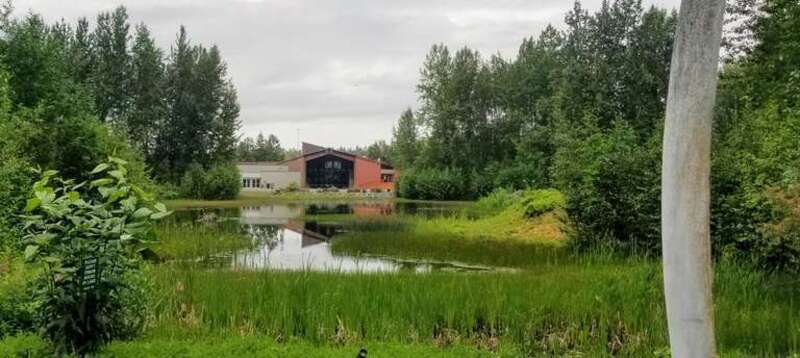 The Alaska Native Heritage Center as seen from across Lake Tiulana. The windows of the Gathering Place can be seen in the distance. A small part of an upright set of whale bones is seen on the right.