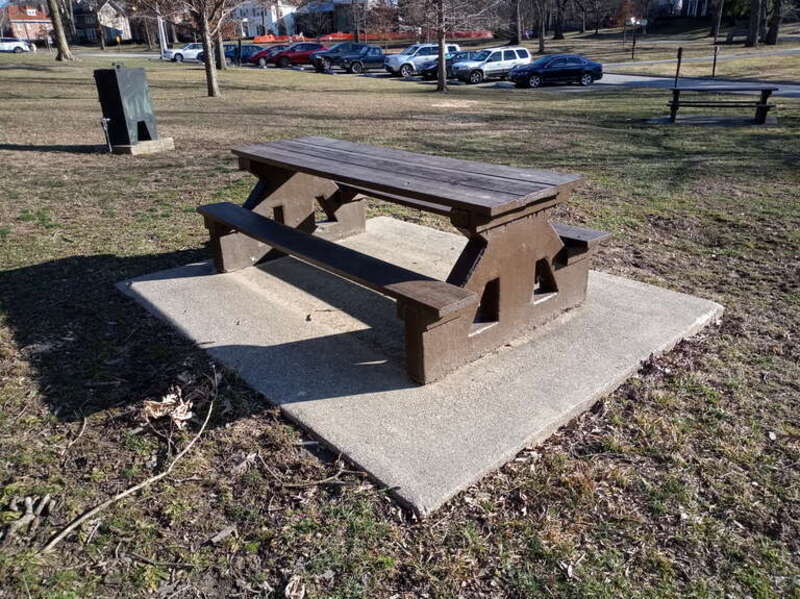 A picnic table of mixed concrete and wood construction, painted a uniform dark brown, on a cement pad at Washington Park in Springfield Illinois.
