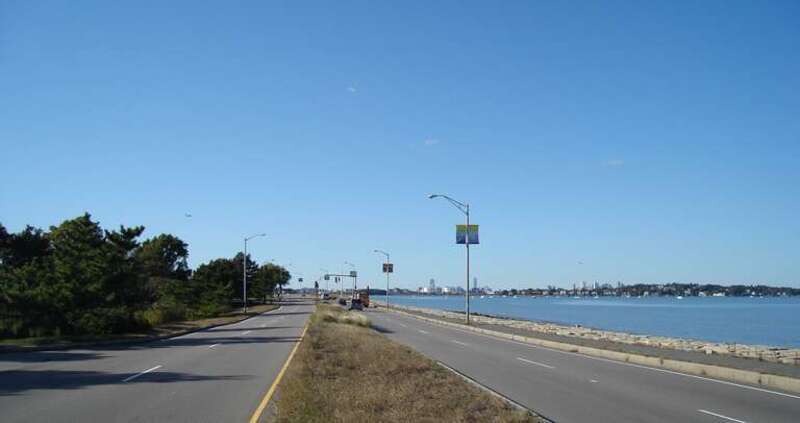 View looking northwest of Quincy Shore Drive in Quincy, Massachusetts with Massachusetts Department of Conservation and Recreation parkland on the left, and the Squantum peninsula and Boston skyline visible center to right beyond the seawall and