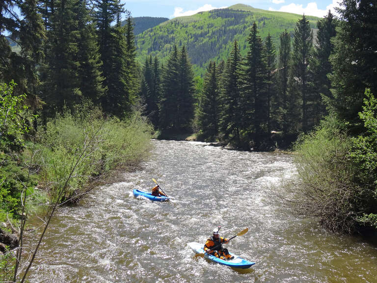 Gore Creek. The view is in Vail, Colorado, looking upstream. Some kayakers are visible in the creek. The picture was taken in early June, 2014, and at that time the creek was swollen with spring runoff from the melting snow.