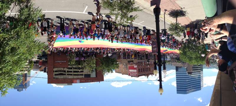 The rainbow flag traveling west on 200 South at the back of the Utah Pride parade in Salt Lake City. People watch from the street, from rooftops, and from a parking structure. Taken using panorama mode on an iPhone 5.