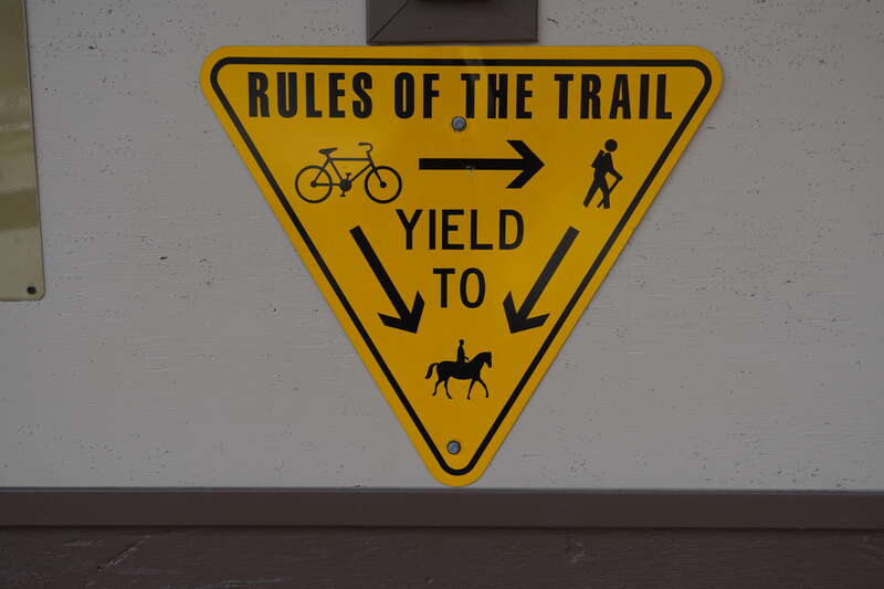 A yellow sign reading 
RULES OF THE TRAIL
YIELD TO

three symbols representing a bike, a hiker, and a horse rider are visible. The sign is telling the viewer that bikers should yield to pedestrians and horses, and pedestrians should yield to horses.