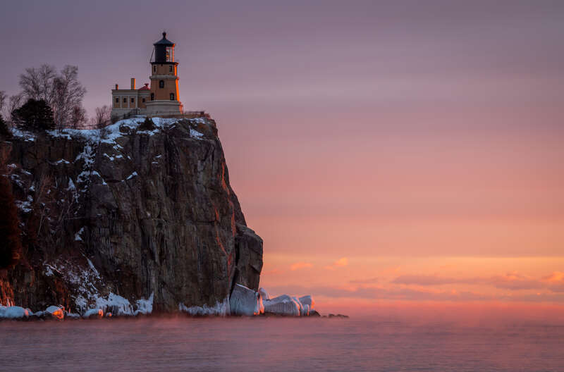 An early morning January photograph of the Split Rock Lighthouse, located near Two Harbors, Minnesota, on the shores of Lake Superior. The image captures the historic lighthouse perched atop a steep cliff, bathed in the soft pastel hues of dawn. The