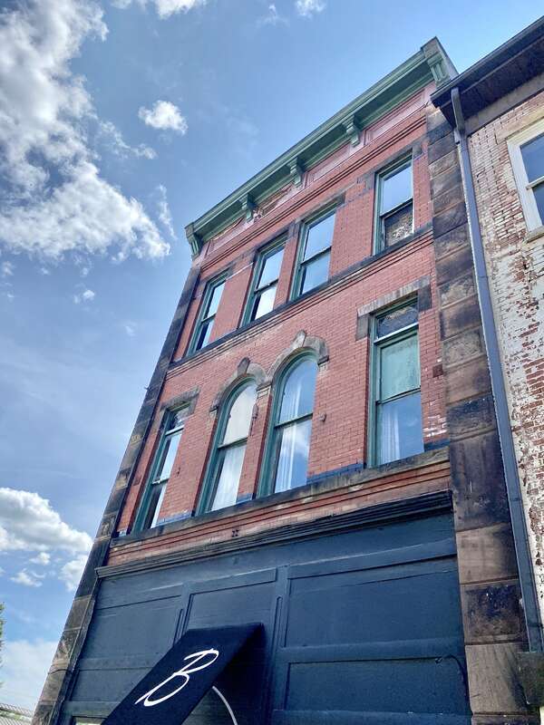 Built in 1875, this Italianate-style building features a red brick exterior, a ghost sign on the south facade, a bracketed cornice, a low-slope rear shed roof surrounded by a parapet, four bays on the second and third floors containing one-over-one
