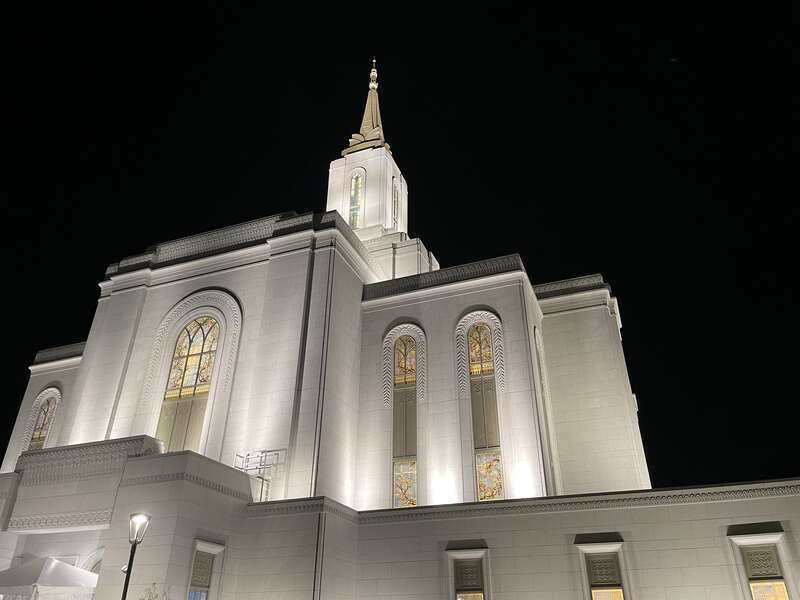 The Orem Utah Temple, seen from below, looking skywards, at night. White parapets stretch tall into the sky, the building has stained glass windows featuring cherry trees. Decorative stonework surrounds the windows with vegetal elements of cherry