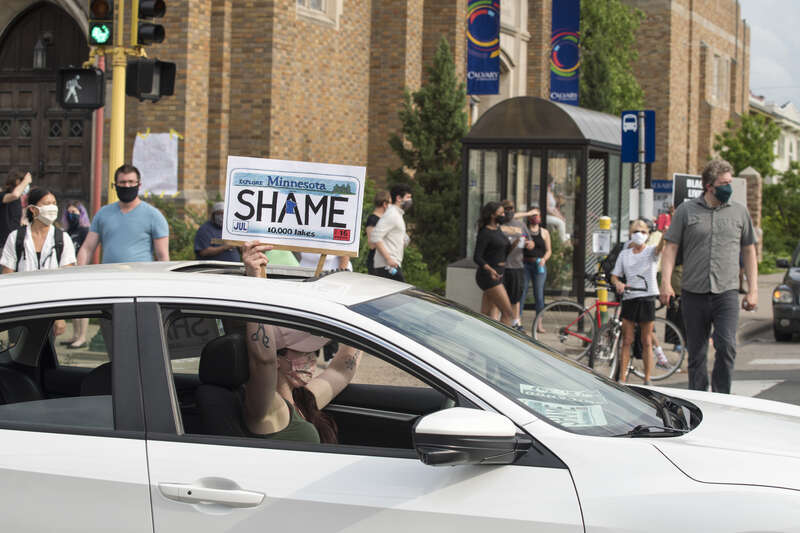 On May 26, 2020, people protested against police violence after the death of George Floyd. A crowd stands on a corner while a protester holds a &quot;Shame&quot; poster out of their car.