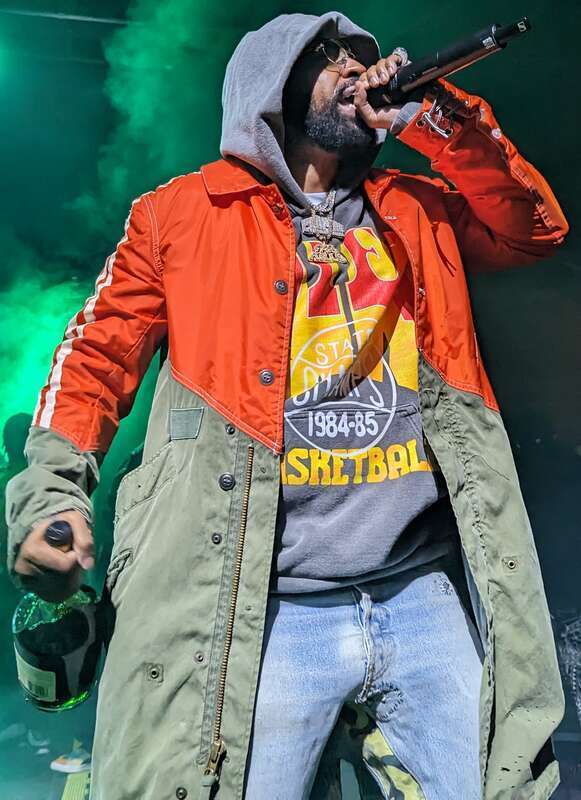 Rahkeim Meyer during a vocal performance with a microphone in his left hand and a bottle of Hennessy Cognac in his right hand while wearing a long two toned orange and light green jacket, with a basketball themed hoodie underneath, and light jeans.