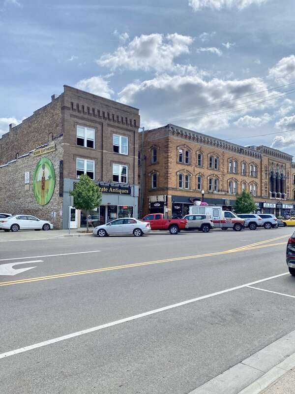 Built around the turn of the 20th Century, this building features a brown brick facade, paired one-over-one windows, stone belt coursing, brick cobbling, a rooftop parapet, a modified first floor retail shopfront, a low-slope shed roof, and an entry
