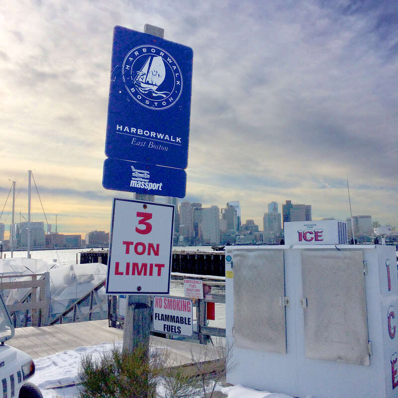 Boston Harborwalk sign, seen from standing at the beginning of the pier at HarborArts, East Boston Shipyard and Marina. Winter sunset over Boston skyline in background.