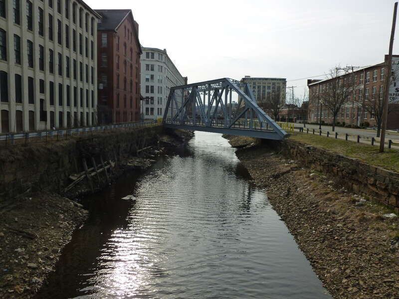 A steel bridge over North Canal, as viewed when looking west fromthe Union Street in Lawrence, Massachusetts. Unused, it is a holdover of Lawrence's beginnings as a trail town.