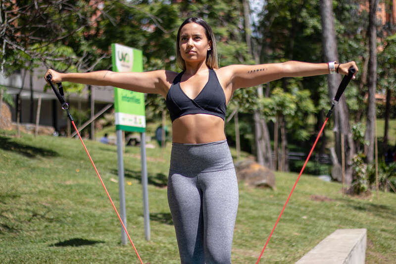 Must attribute with link to: www.ptpioneer.com
Image of a girl Doing Lateral raises with red elastic bands in a park Facing the camera.