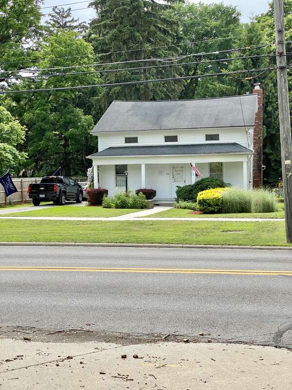 Built in the mid-19th Century, this one-and-a-half-story Greek Revival-style house features a wooden exterior with three small attic windows on the front facade, six-over-six double-hung windows, a mid-20th Century front door, a porch with square